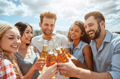Cheers to friends! Group of happy young people are clinking bottles with beer and smiling while standing on the roof. Barbecue concept.