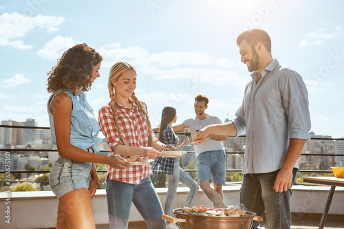Delicious food for best friends. Young and cheerful man is putting hot barbecued meat on the plates of his female friends while standing on the roof. Barbecue concept.