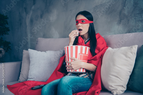 Close up photo beautiful she her superpower lady costume hold popcorn container controller change channels scary movie brave but fear wear red eye mask mantle sit sofa couch divan house indoors