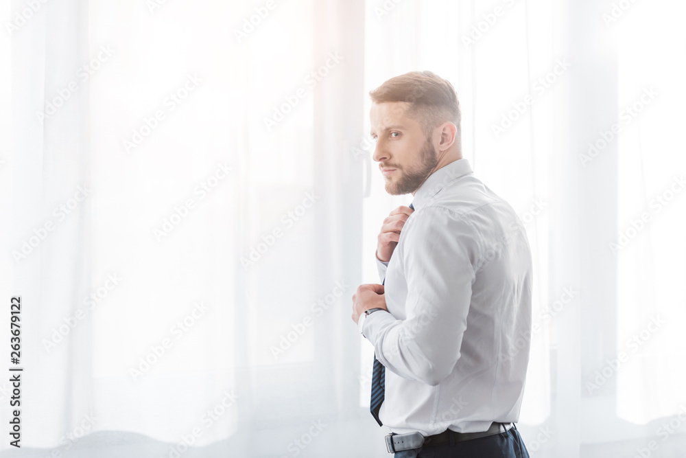 Fototapeta premium handsome bearded man touching tie while standing at home