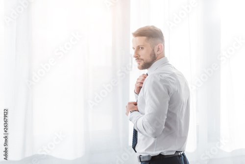 handsome bearded man touching tie while standing at home