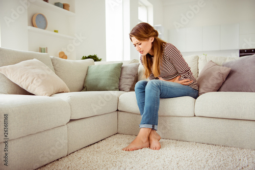 Portrait of upset disappointed lady sit divan touch tummy had indigestion food poisoning hurt injury suffering dressed in denim outfit striped shirts close eyes long curly wavy hair in apartment