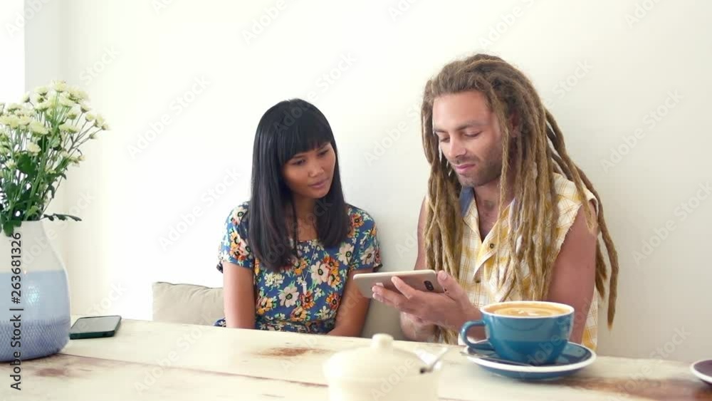 Black hair asian girl discuss with caucasian dreadlock man sitting at the table in coffee shop and watching something on smartphone Black hair asian girl discuss with caucasian dreadlock man sitting at the table in coffee shop and watching something on smartphone