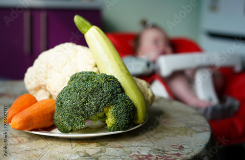 a plate of vegetables - broccoli, zucchini, carrots and cauliflower. Little baby on the background.