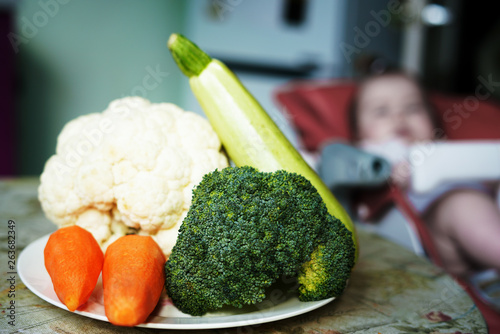 a plate of vegetables - broccoli, zucchini, carrots and cauliflower. Little baby on the background.