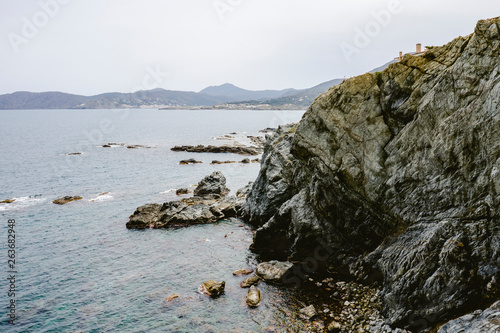 Coastal landscape with rocks and stones next to the beach, the sea and a coastal village with clear blue sky.