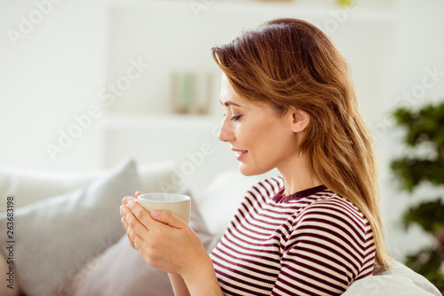 Close up side profile photo beautiful amazing she her lady imaginary flight calm kindhearted hold hot beverage hands arms wear striped pullover clothes sit comfort divan flat house living room indoors