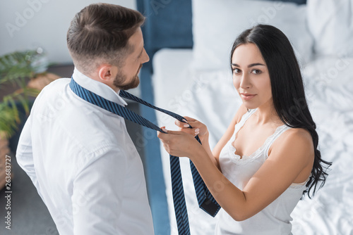 overhead view of beautiful woman tying tie of bearded man in bedroom