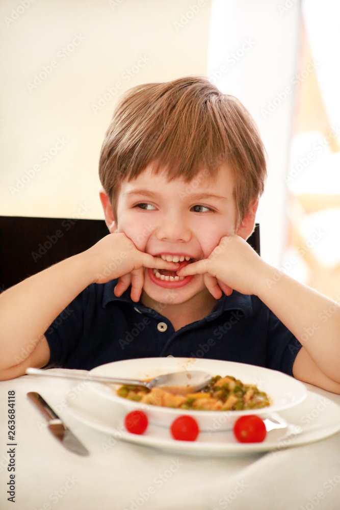 Hungry child sitting in chair at table in kitchen and eating with spoon of cooked peas with tomato. Kids meal and healthy diet food concept. Happy and cute little boy enjoying in good lunch at home. Hungry child sitting in chair at table in kitchen and eating with spoon of cooked peas with tomato. Kids meal and healthy diet food concept. Happy and cute little boy enjoying in good lunch at home.