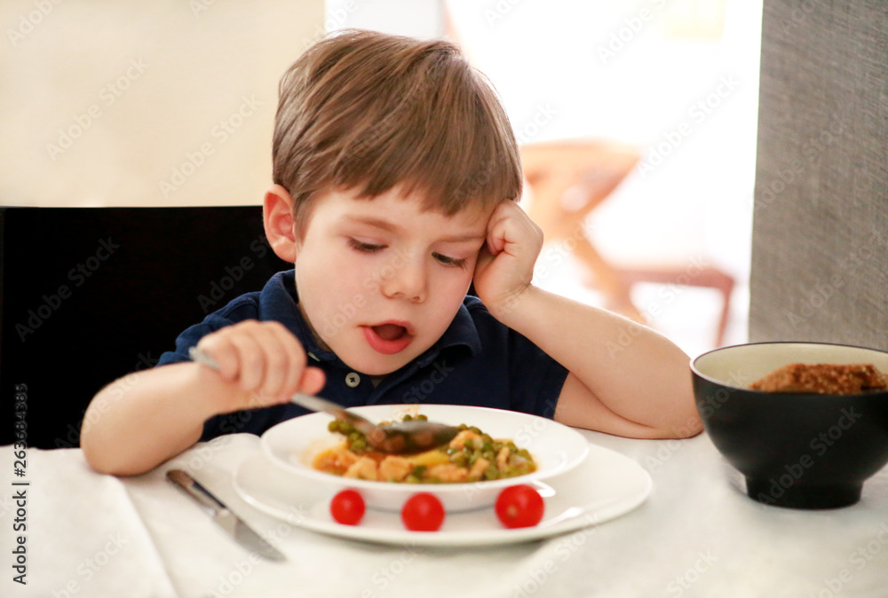 Hungry child sitting in chair at table in kitchen and eating with spoon of cooked peas with tomato. Kids meal and healthy diet food concept. Happy and cute little boy enjoying in good lunch at home. Hungry child sitting in chair at table in kitchen and eating with spoon of cooked peas with tomato. Kids meal and healthy diet food concept. Happy and cute little boy enjoying in good lunch at home.