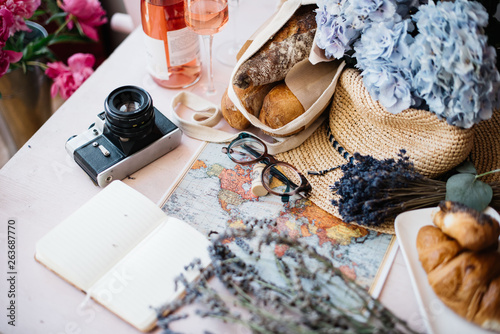 summer travel flat lay: a glass of rose wine, blue hydrangea flower, wicker hat, croissant, world map, a bag of baguettes, dry lavender