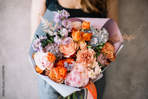 Very nice young woman holding beautiful blossoming flower bouquet of fresh peony, roses, carnations, mattiola, eucalyptus in orange and lavender colours on the grey wall background