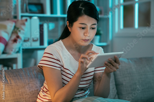 elegant asian woman using tablet computer for sharing social media sitting on sofa at home at night. girl in dark living room in midnight browsing website online internet. young people technology