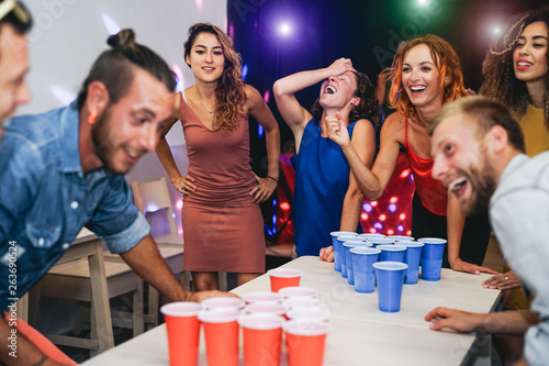 Happy friends playing beer pong in a cocktail bar - Young millennial people having fun doing party alcohol games at night pub - Friendship and youth lifestyle nightlife concept