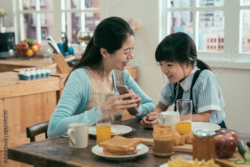 Funny mom and lovely child are having fun with phone. young asian housewife in apron and cute daughter in uniform sitting at morning kitchen table with tasty healthy meal breakfast. kid interested.