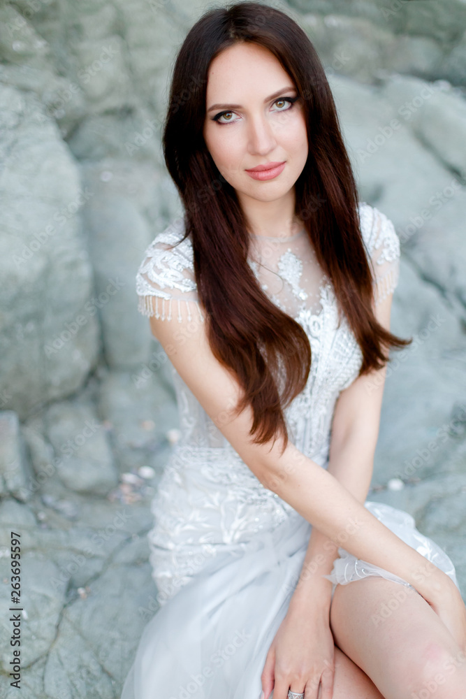 Young beautiful brunette bride in light grey chiffon wedding dress embroidered with beads posing on a rocky beach near the sea. Young beautiful brunette bride in light grey chiffon wedding dress embroidered with beads posing on a rocky beach near the sea.
