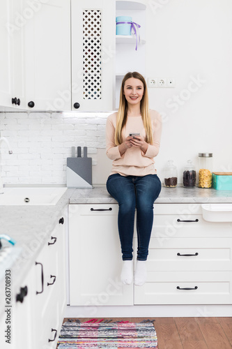 Adorable nice charming cheerful beautiful trendy stylish girl chatting on mobile sitting on table countertop in modern light white kitchen