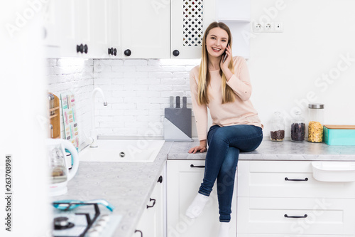 Adorable nice charming cheerful beautiful trendy stylish girl chatting on mobile sitting on table countertop in modern light white kitchen