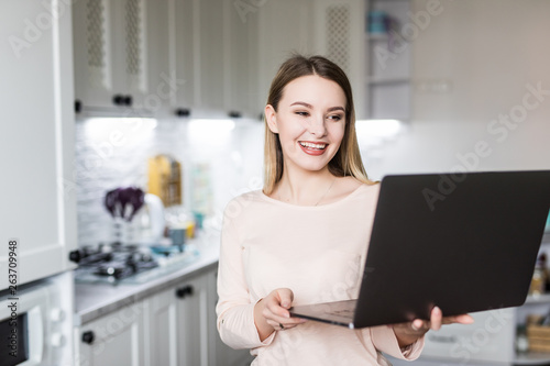 Happy female looking at laptop while standing in kitchen in morning.