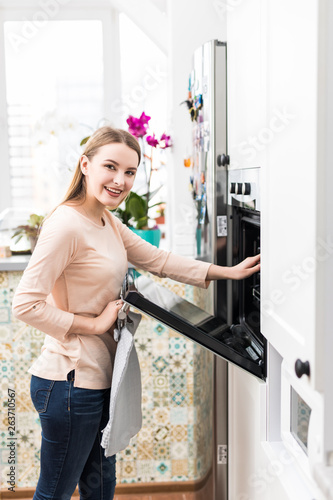 Beautiful young woman opening door of oven with baked buns in kitchen