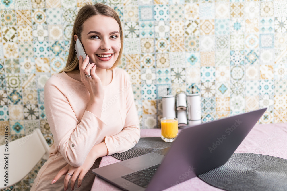 Young woman sits at the kitchen table using a laptop and talking on a cell phone Young woman sits at the kitchen table using a laptop and talking on a cell phone
