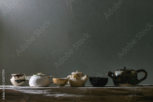 Variety of craft handmade ceramic teapots and cups for tea ceremony standing on old wooden shelf in dark room.