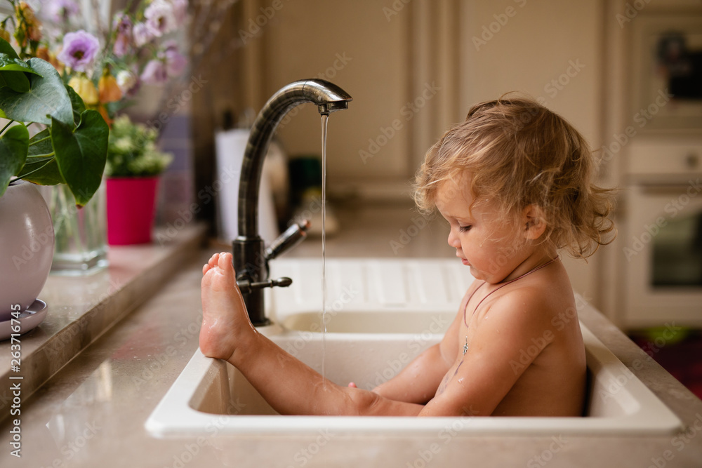 Baby taking bath in kitchen sink. Child playing with foam and soap bubbles in sunny bathroom with window. Little boy bathing. Water fun for kids. Hygiene and skin care for children - obrazy, fototapety, plakaty 