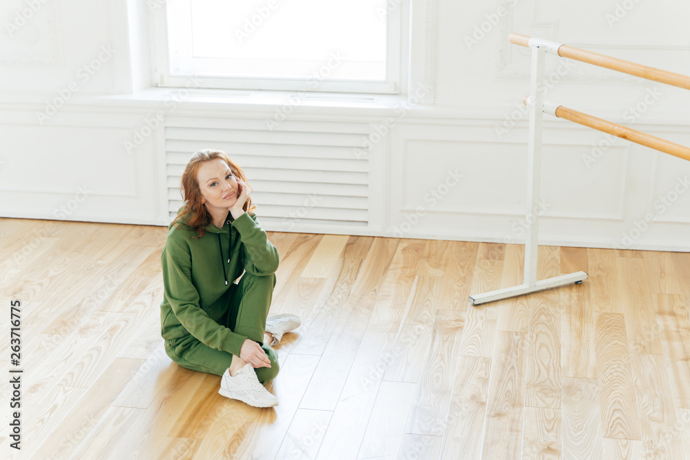 Photo of professional female dancer has rest after training, poses on floor near ballet barre, has ballet classes, wears green sweatsuit, sportshoes, likes being active. People, fitness and exercising Photo of professional female dancer has rest after training, poses on floor near ballet barre, has ballet classes, wears green sweatsuit, sportshoes, likes being active. People, fitness and exercising