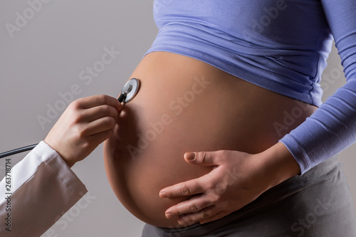Image of doctor examining a pregnant woman with stethoscope on gray background.