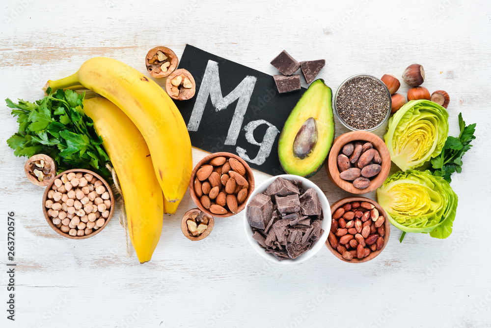 Foods containing natural magnesium. Mg: Chocolate, banana, cocoa, nuts, avocados, broccoli, almonds. Top view. On a white wooden background. - obrazy, fototapety, plakaty 