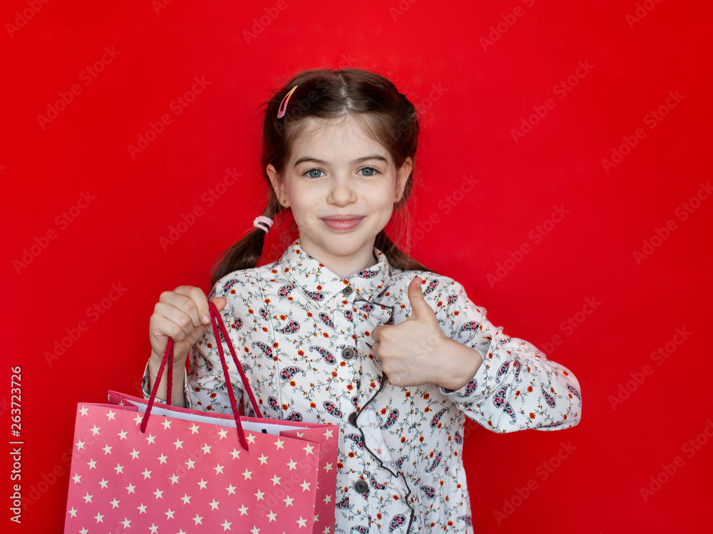 smiling young girl holding a gift bag in her hands and showing thumbs up on a red background, concept of advertising, sales, discounts smiling young girl holding a gift bag in her hands and showing thumbs up on a red background, concept of advertising, sales, discounts