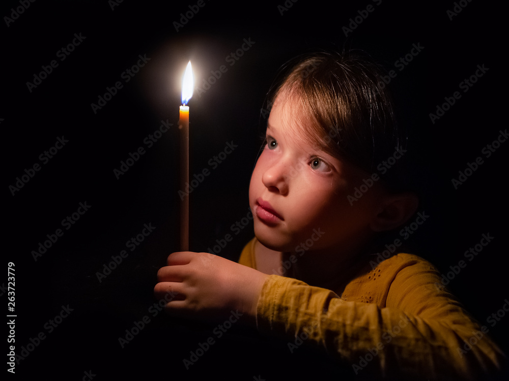 little girl prays and looks at a burning candle in the dark