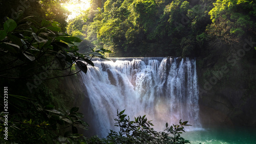 Beautiful scenery of natural waterfall inside the forest with warm sunlight effect shining through the inside. Foreground have tree branches. Selected focus.