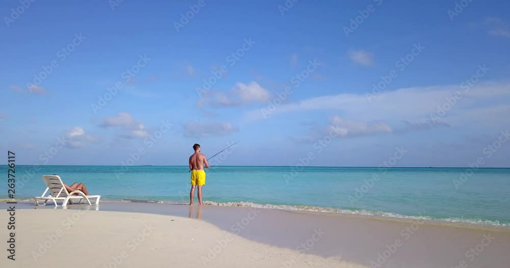 Athletic man fishing in beautiful while girl relaxes on beach chair, pull back shot 4k Athletic man fishing in beautiful while girl relaxes on beach chair, pull back shot 4k