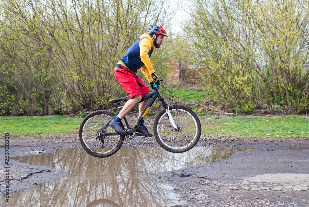 A cyclist in red shorts and a yellow jacket riding a bicycle on the rear wheel through a puddle.