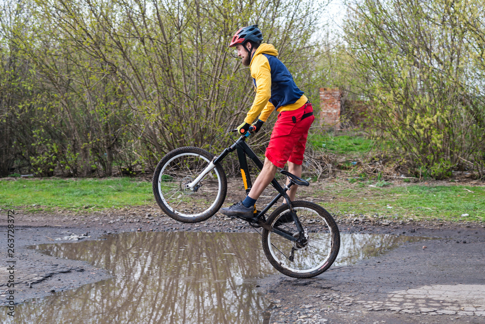 A cyclist in red shorts and a yellow jacket riding a bicycle on the rear wheel through a puddle.