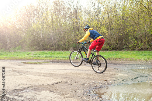 A cyclist in red shorts and a yellow jacket riding a bicycle on the rear wheel through a puddle.