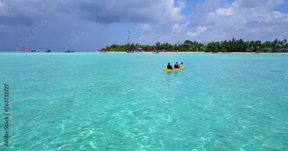Kayakers slowly paddle their yellow kayak through clear shallow coastal water in the Maldives to a palm covered island as gray clouds gather in background. Aerial, follow, 4k. Kayakers slowly paddle their yellow kayak through clear shallow coastal water in the Maldives to a palm covered island as gray clouds gather in background. Aerial, follow, 4k.