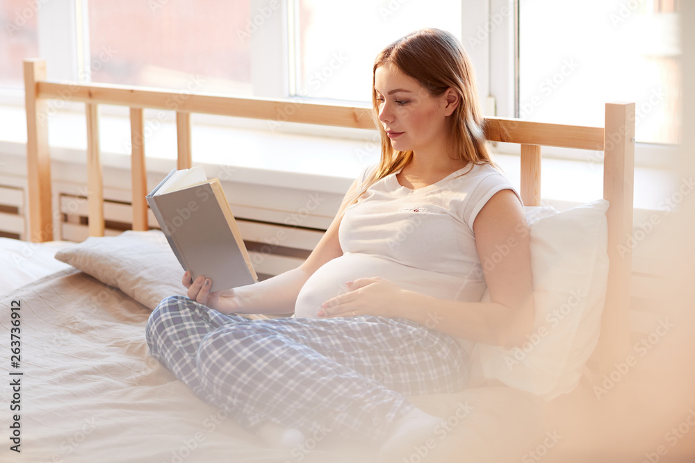 Full length portrait of young pregnant woman lying on bed and reading book in warm sunlight, copy space Full length portrait of young pregnant woman lying on bed and reading book in warm sunlight, copy space