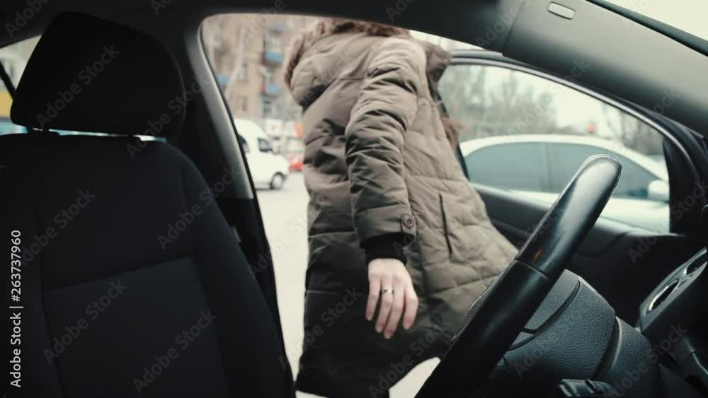 A young girl, a driver, a brunette with long hair, in a long khaki jacket, comes out of her car. View from the car. Slow motion A young girl, a driver, a brunette with long hair, in a long khaki jacket, comes out of her car. View from the car. Slow motion