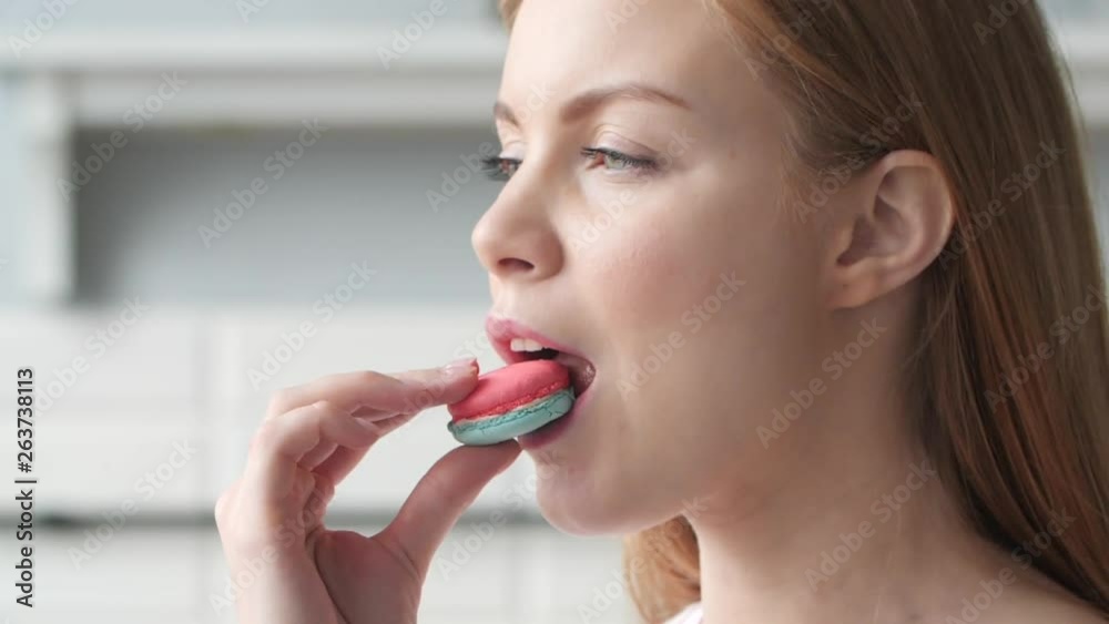 Brunette girl in nude t-shirt standing on kitchen and eating cookie. Young beautiful girl in pijama enjoying sweets on breakfast. Happy cute woman holding delicious red macaron in hand and smiling. Stock Video | Adobe Stock
