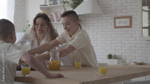 Portrait young pretty mother bakes pies for her four teen sons in white shirts waiting for the food sitting at the table in the kitchen. Big friendly noizy family having breakfast together photo