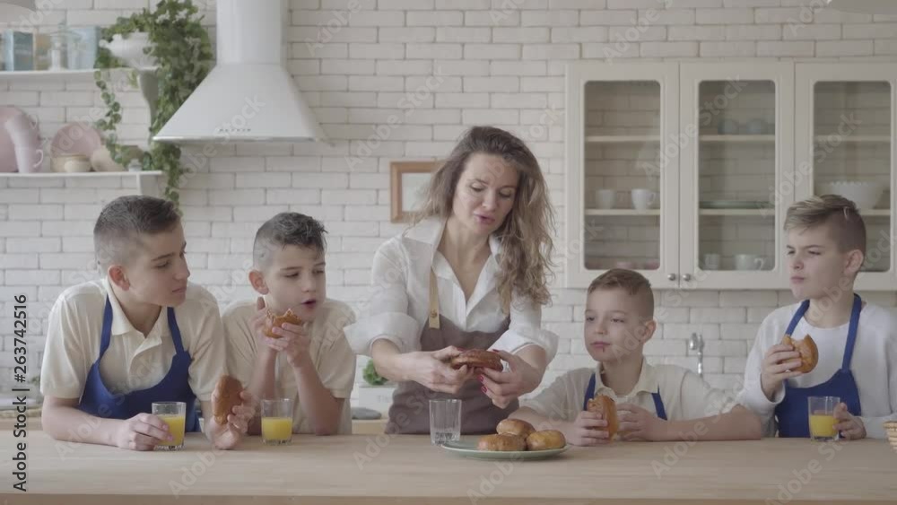 Portrait attractive happy woman and her four teen son eating pies and drinking orange juice in the kitcken standing at the table. Everybody wearing aprons. Kids helping mother to cook food