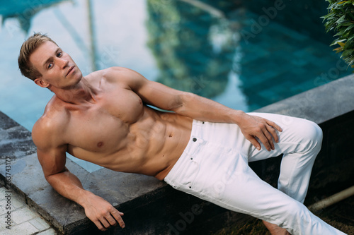 Sexy fitness man in white pants with sporty body sits near swimming pool