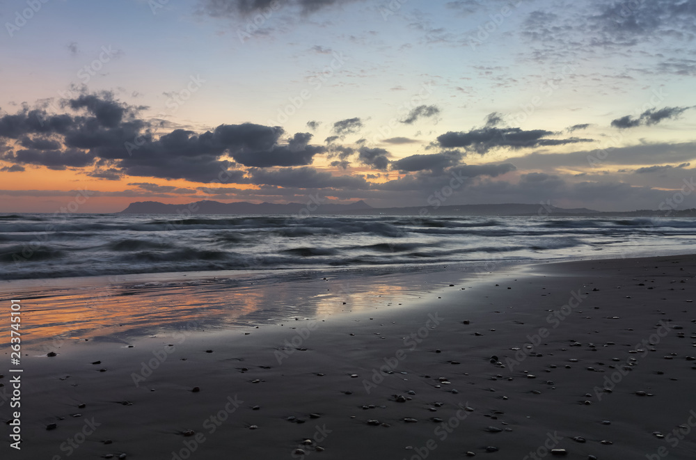 Location place Agia Marina Beach, island Crete, Greece. Sea coast spangled by rocks, the sunrise is reflecting on the wet sand. The mountains in haze on the horizon. Storm sea with high waves.