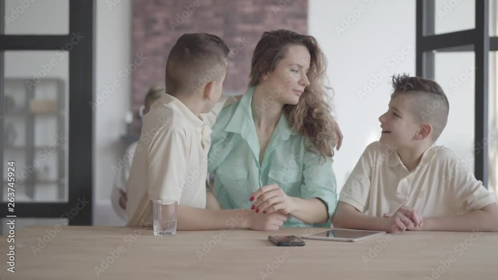 Sons hug tired mom standing at the table indoors. Brotherhood. Family relationship. Sons hug tired mom standing at the table indoors. Brotherhood. Family relationship.