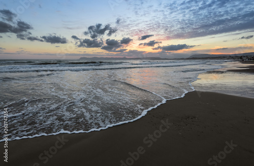 High waves with foam spread on the sand on the coast. The light of unbelievable sunset is reflecting on the sea. Pre dawn time. Beautiful enlighten sky with clouds. Mountains. Romantic relax place.