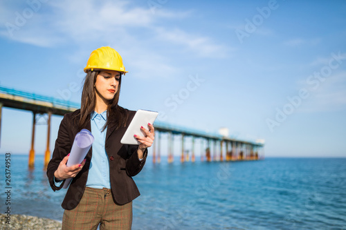 Giovane ragazza ingegnere con tablet in mano, progetto arrotolato di carta è sulla costa di fronte al cantiere di un approdo navale in Oceano. photo
