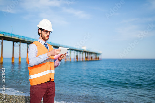 Giovane ragazzo ingegnere con gilet giallo, caschetto bianco e tablet in mano, sta osservando la costruzione di un ponte in mare aperto, dalla costa photo