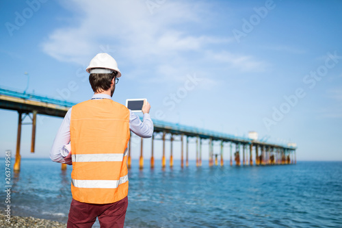Giovane ragazzo ingegnere con gilet giallo, caschetto bianco e tablet in mano, sta osservando la costruzione di un ponte in mare aperto, dalla costa photo